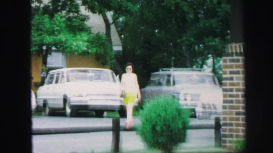 Young woman in Lynbrook, New York, walks toward the camera in bright shorts, a white sleeveless top, and sunglasses near parked cars.