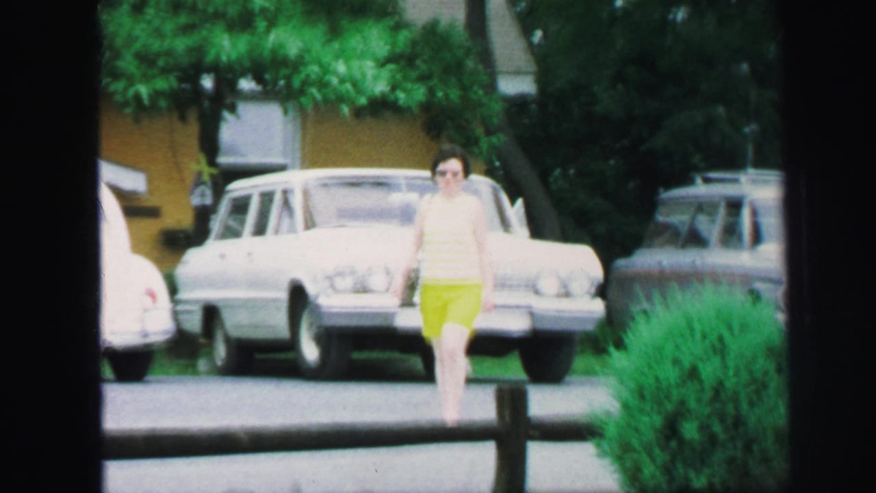 The woman continues walking toward the camera in 1971 Lynbrook, with a white station wagon and leafy residential background behind her.