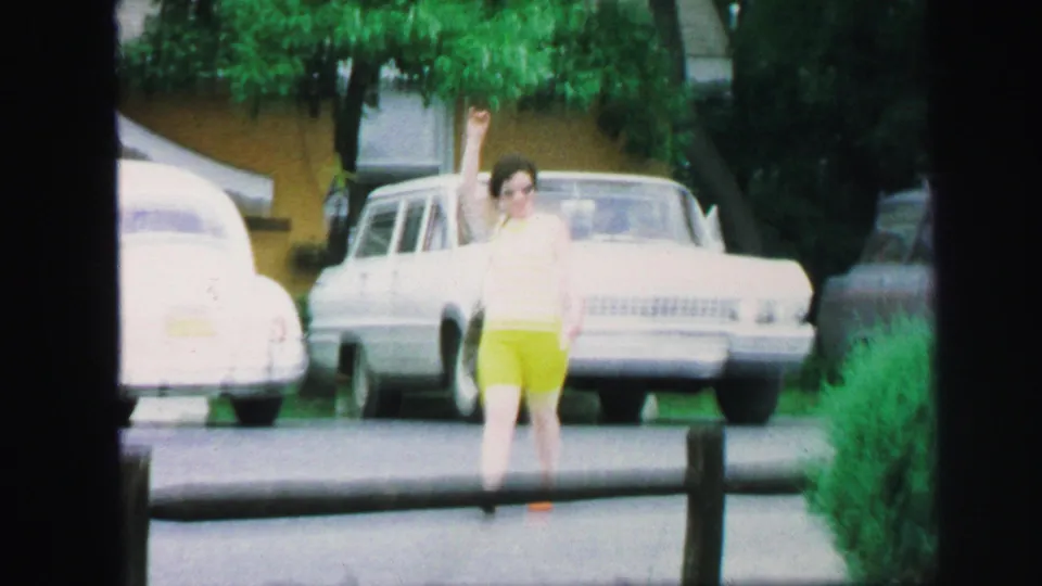 In a Lynbrook street scene, the woman raises her arm in a wave while framed by parked older cars, greenery, and a low rail.