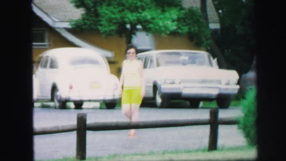 The woman walks along the Lynbrook street after waving, with a white Volkswagen Beetle, larger parked car, trees, and shrubs visible.