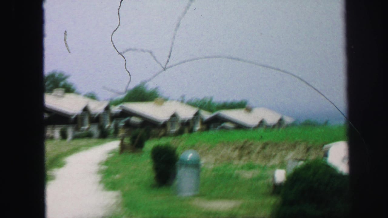 Cabins and a pale walkway sit beside grass, shrubs, and a small trash can.