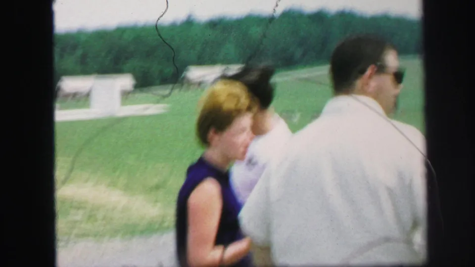 Adults in casual clothing walk through Lynbrook resort grounds, with cabins, lawn, and wooded hills behind them.