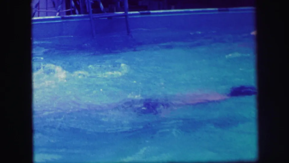 1971 Lynbrook home-movie frame showing a swimmer gliding through the backyard pool under blue-purple shifted film color.