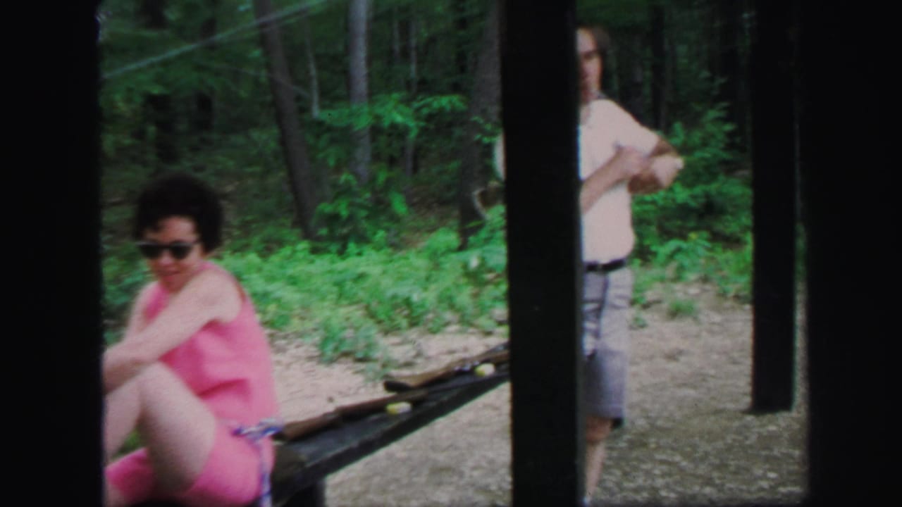 Woman in pink swimsuit sits on a bench near rifles in a wooded range area.