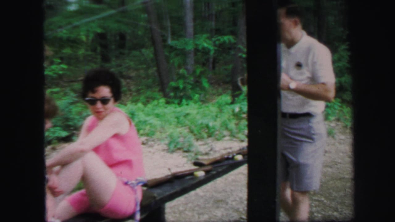 1971 Lynbrook home-movie frame with a seated woman, nearby rifles, and a man standing beside a wooded shooting range.