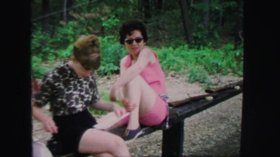 1971 Lynbrook home-movie frame showing two women seated near rifles and a bench at a wooded outdoor range.