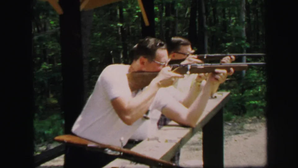 1971 Lynbrook home-movie frame showing two men aiming rifles from a wooden shooting station surrounded by trees.