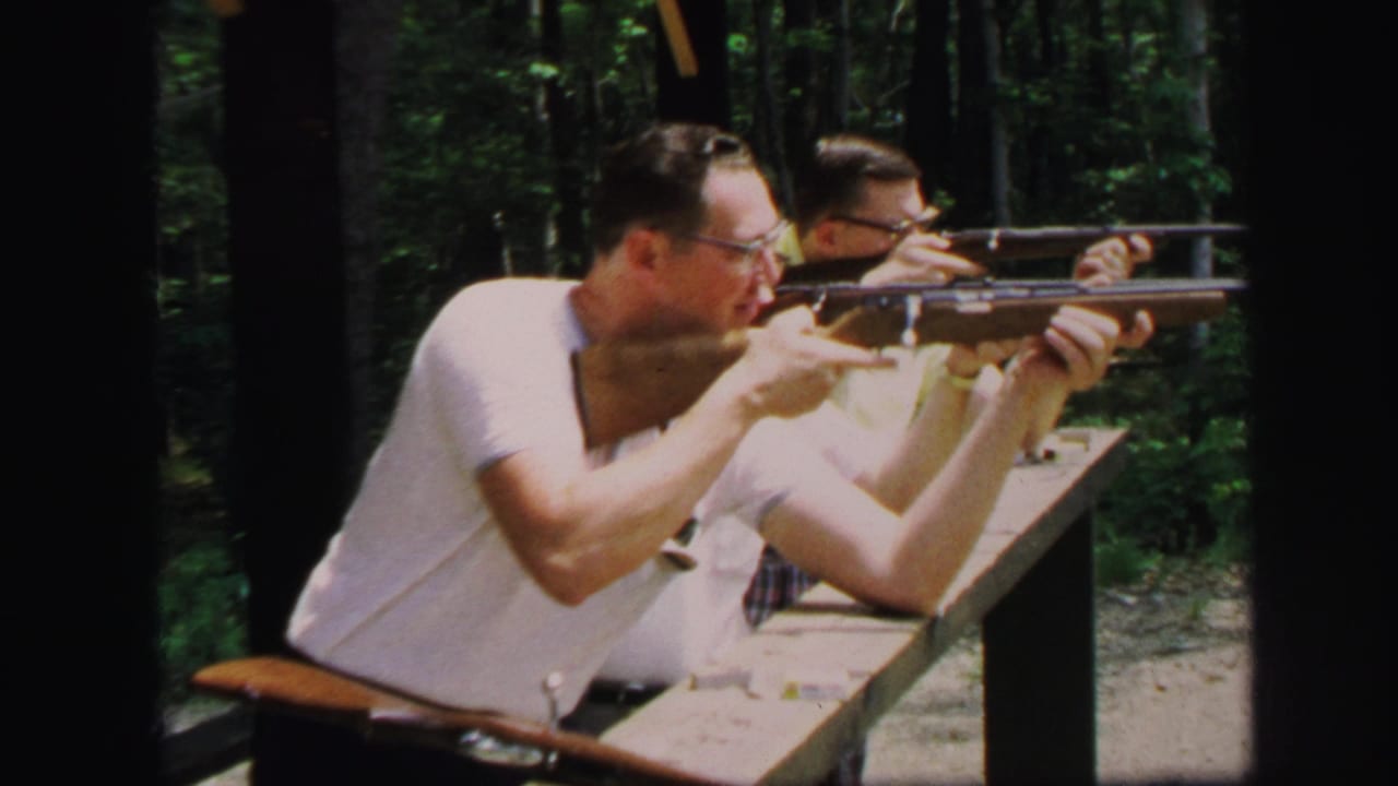 In Lynbrook, New York archival 8mm footage, adult men in light shirts aim rifles from a wooden bench in a wooded outdoor range.