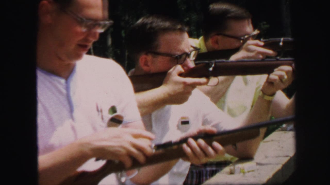 Adult men handle and aim rifles at an outdoor shooting range, with one man in a white shirt prominent in the foreground.