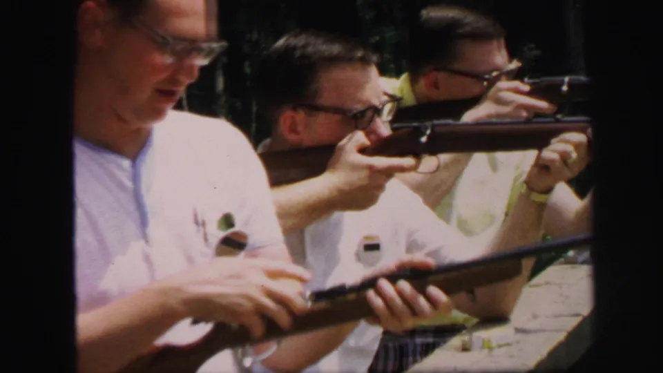 Adult men handle and aim rifles at an outdoor shooting range, with one man in a white shirt prominent in the foreground.