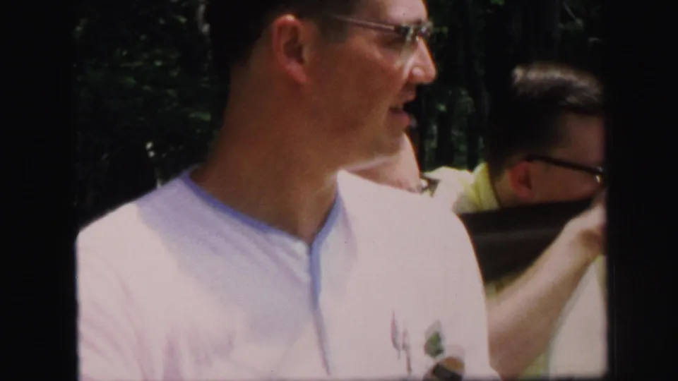 A close 8mm frame shows a man in glasses near other adult men aiming rifles at a wooded outdoor range in Lynbrook.