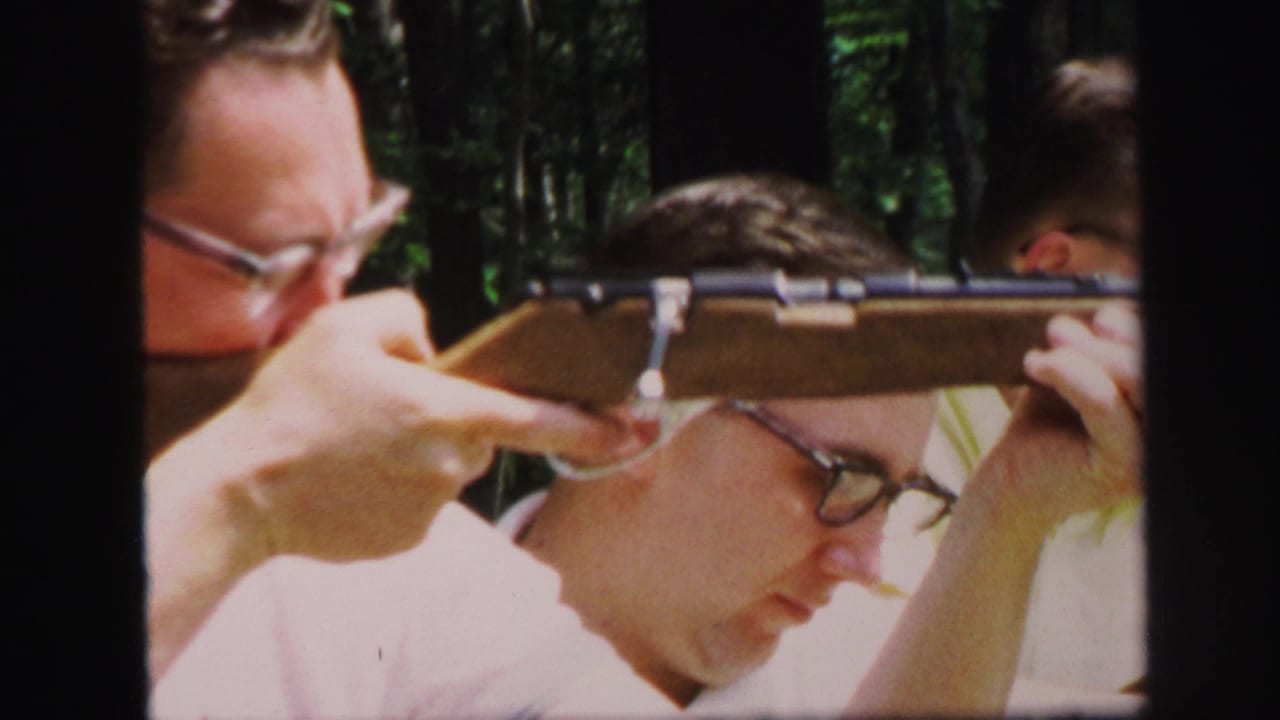1971 Lynbrook frame of a man aiming a long gun at an outdoor range, with other adults close behind in a wooded setting.
