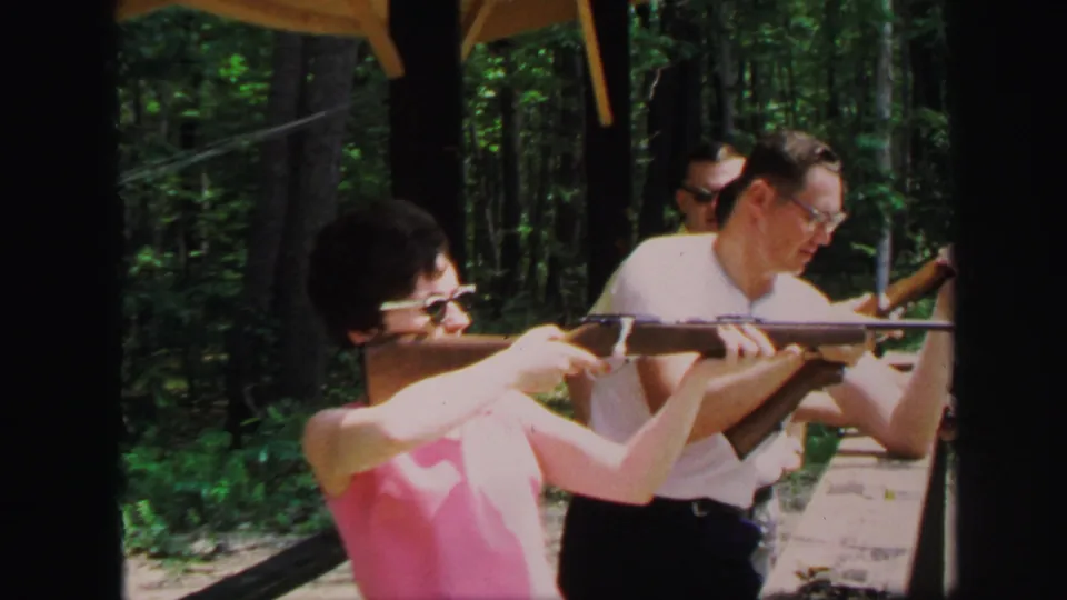 1971 Lynbrook home-movie frame showing a woman in a pink sleeveless top aiming a long gun while adults stand nearby.