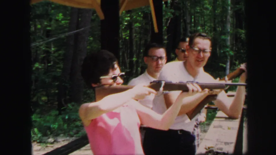 1971 Lynbrook frame of a woman aiming a long gun under a wooden shelter, with men watching in a wooded shooting-range setting.