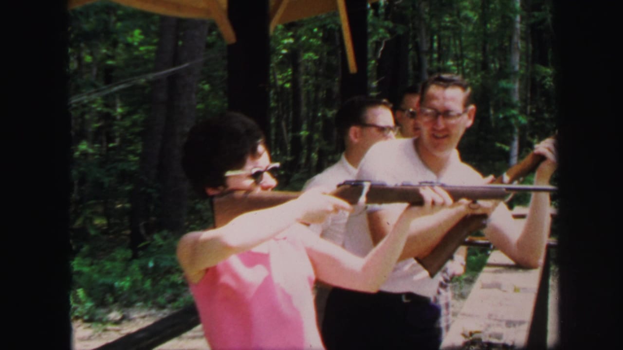 Woman in sunglasses aims a rifle while men stand behind her at a wooded shooting range.