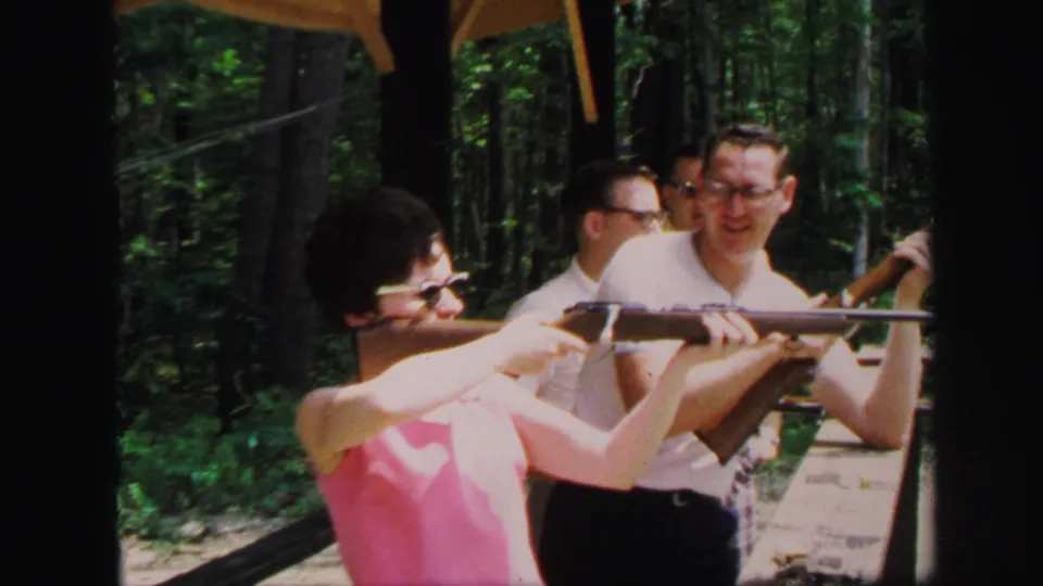 In 1971 Lynbrook, New York footage, a woman in a pink sleeveless top aims a rifle as men observe at an outdoor shooting station.