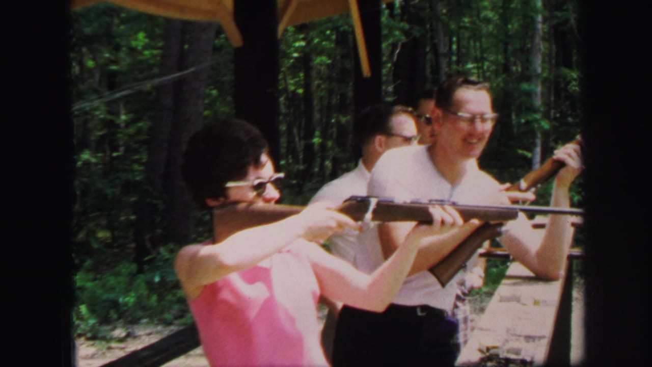A woman aims a long rifle at an outdoor range while a man in a white shirt smiles beside her in 1971 Lynbrook home-movie footage.