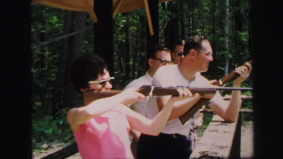 The woman maintains her stance with the rifle as men watch from behind at a wooded Lynbrook shooting range in 1971 home-movie footage.