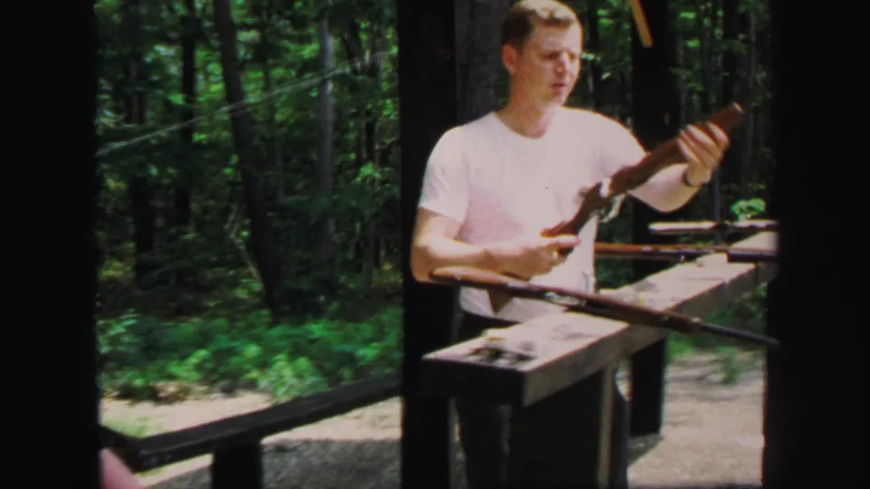 A man in a white shirt holds a rifle near a wooden shooting bench at the wooded range, consistent with the 1971 Lynbrook clip metadata.