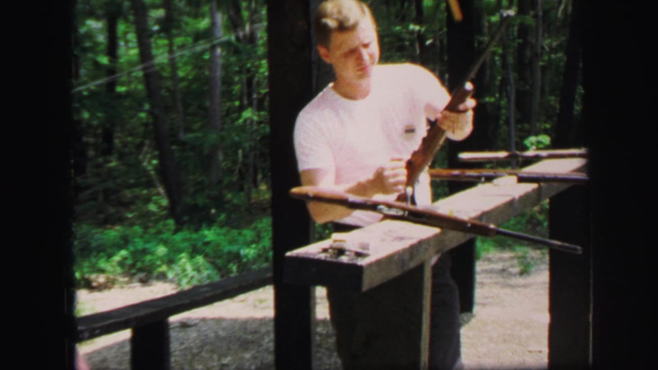 Young man in a white T-shirt holds a rifle at a wooded outdoor shooting bench.