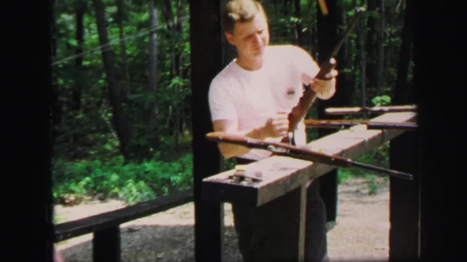 In 1971 Lynbrook, New York footage, a young man stands at a wooden range station while lifting or inspecting a rifle.
