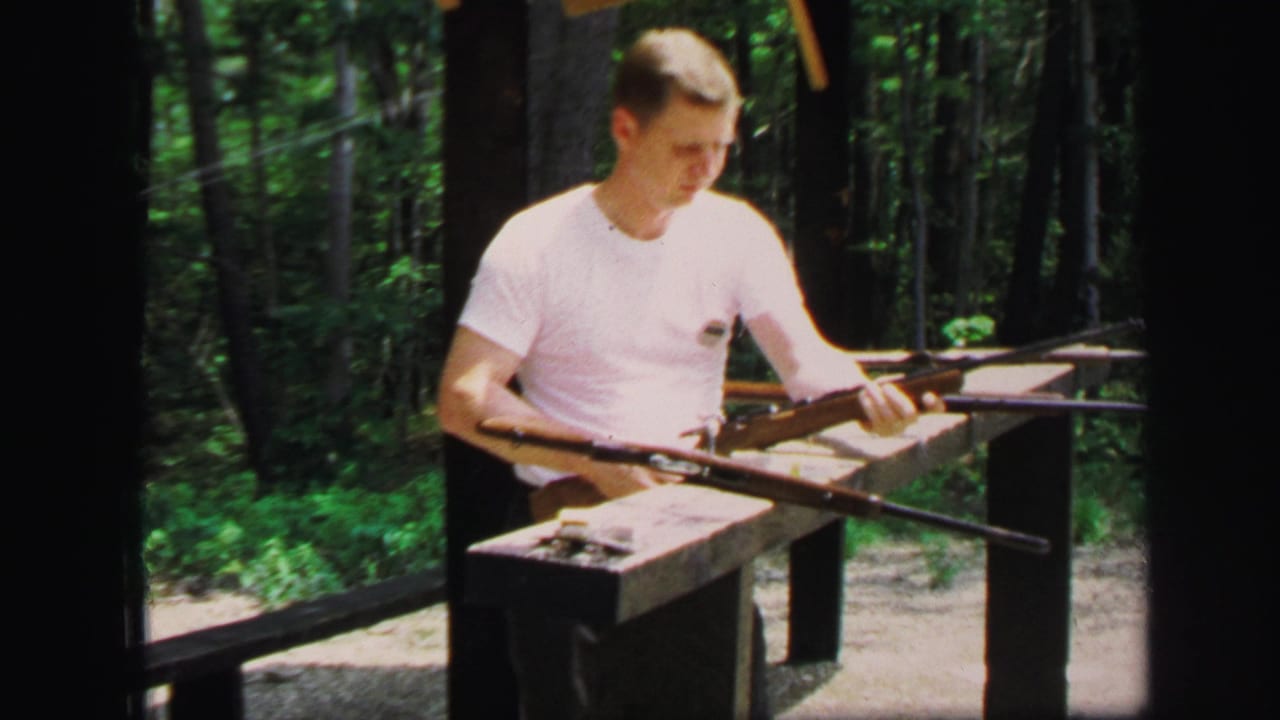 The man at the Lynbrook outdoor range looks down toward a rifle positioned across the bench, with woods behind him.