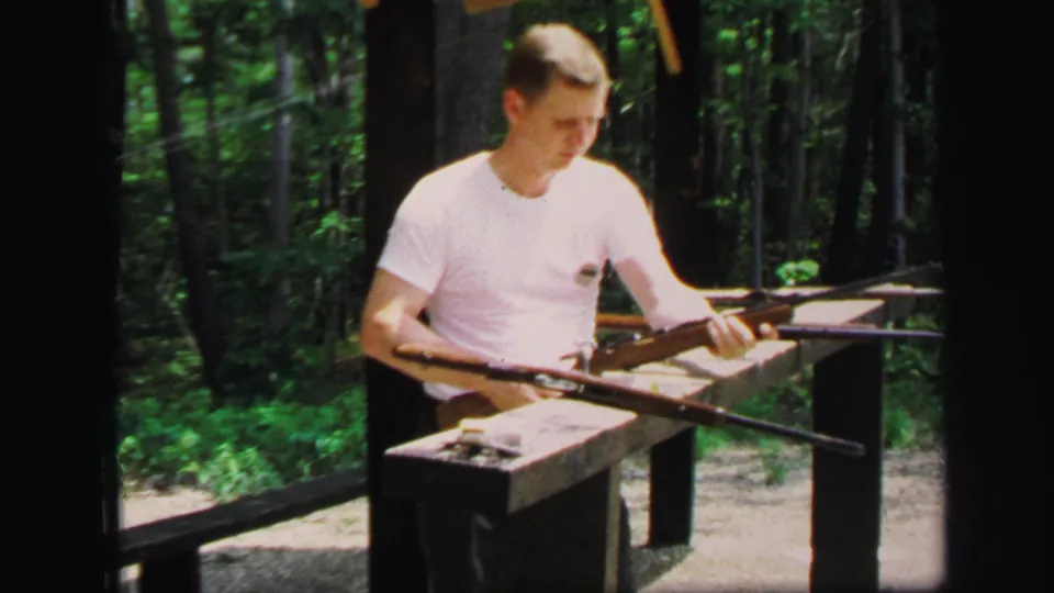 The man at the Lynbrook outdoor range looks down toward a rifle positioned across the bench, with woods behind him.