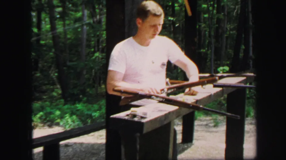 The young man reaches down toward the firearm on the range station, surrounded by trees in the 1971 home-movie scene.