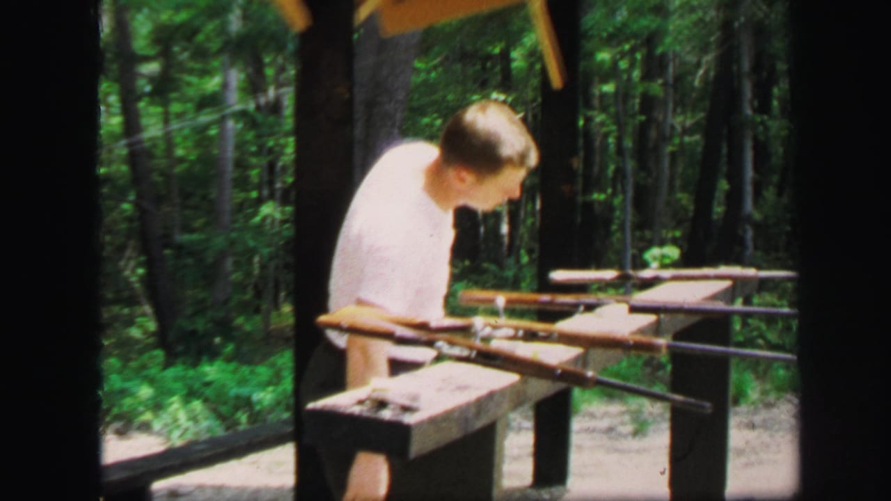 Man in a white shirt stands at a wooden shooting bench with rifles in a wooded range.
