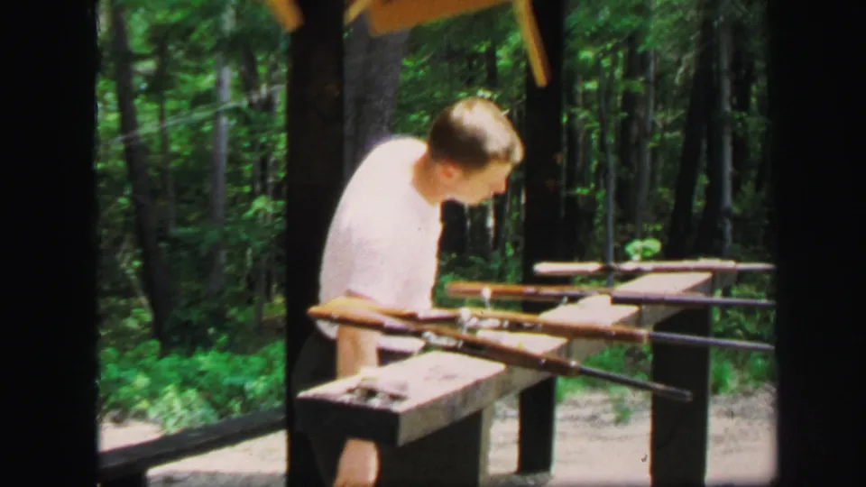 In 1971 Lynbrook, New York footage, a man in a white shirt stands near rifles on a wooden bench at a wooded gun range.