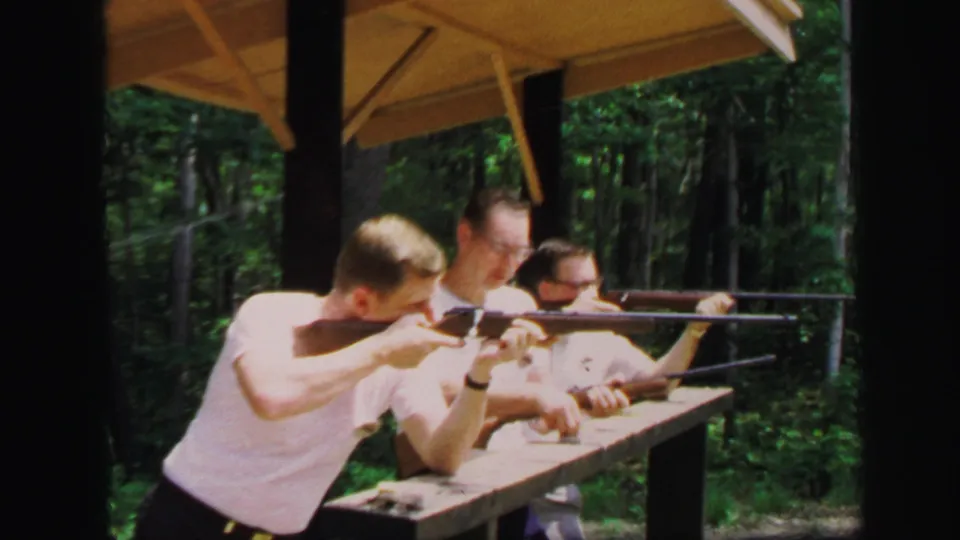 Adult men lean into their rifles at a wooded gun range, with a wooden bench and shelter roof visible in 1971 Lynbrook footage.