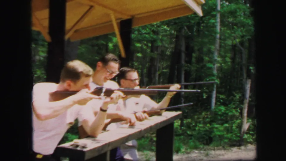 Three adult men handle rifles at a wooden shooting bench beneath a yellow shelter roof in 1971 Lynbrook, New York footage.