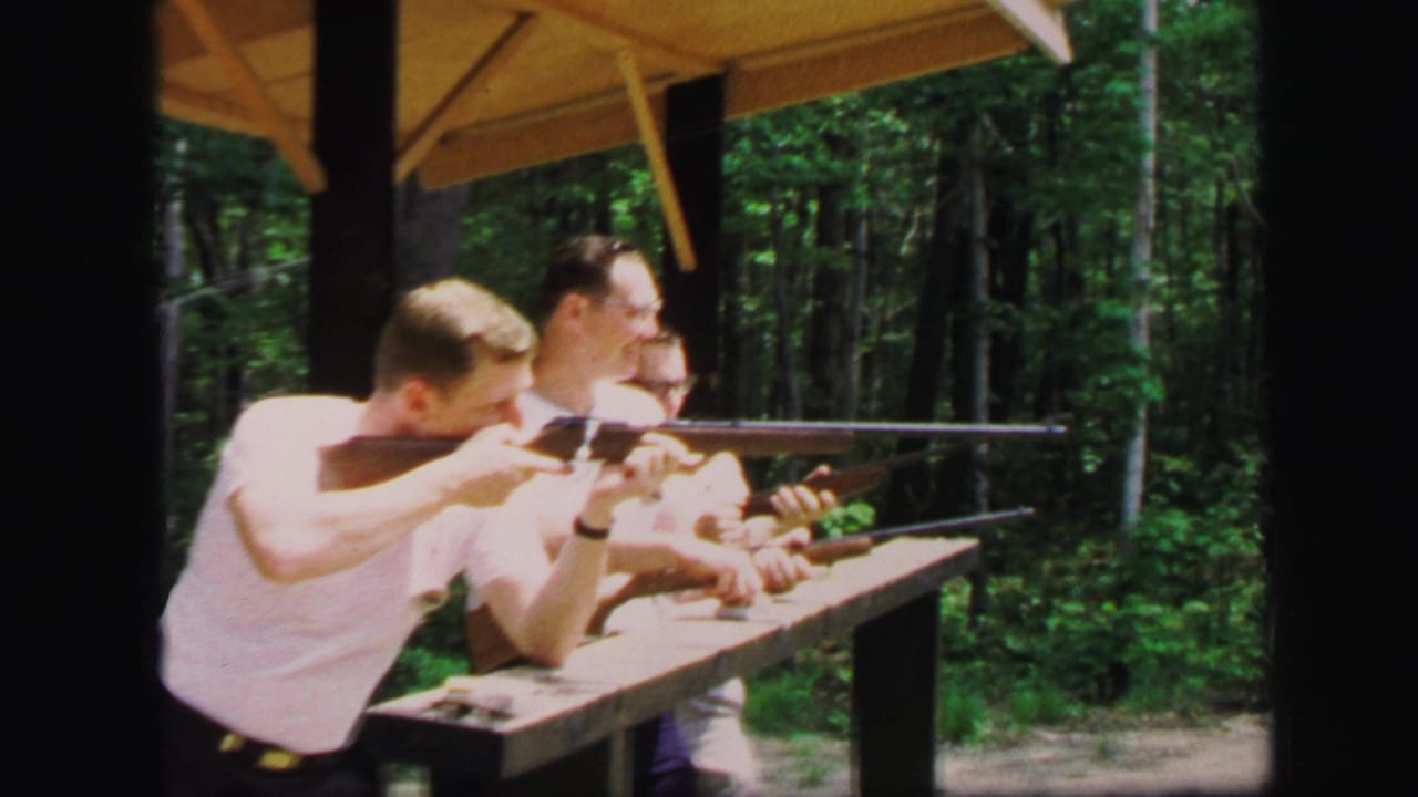 Three young men at a wooden shooting bench aim rifles under a shelter beside trees.