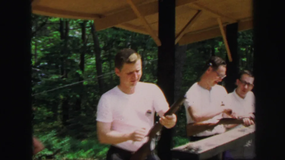 At the 1971 Lynbrook outdoor range, one young man stands with a rifle as two others prepare rifles at the bench.