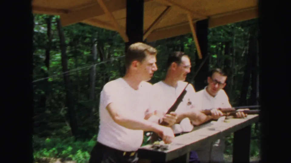 Three young men in 1971 Lynbrook stand at a covered shooting bench with rifles and forest visible behind them.