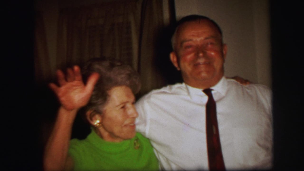 In the supplied 1971 Lynbrook home-movie clip, an older woman in green waves while standing beside a smiling man indoors.