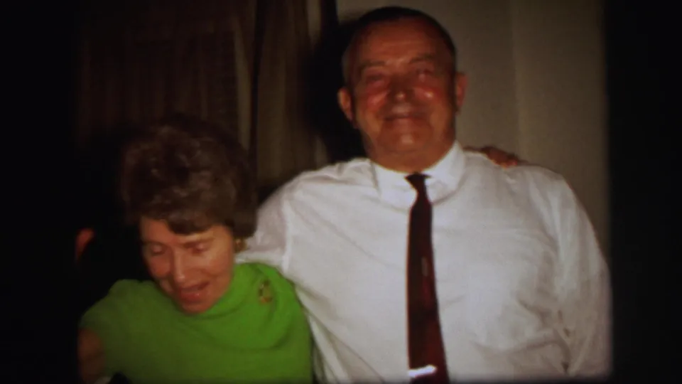 The woman in green looks down as the man in a white shirt and dark tie smiles in the Lynbrook indoor home-movie scene.