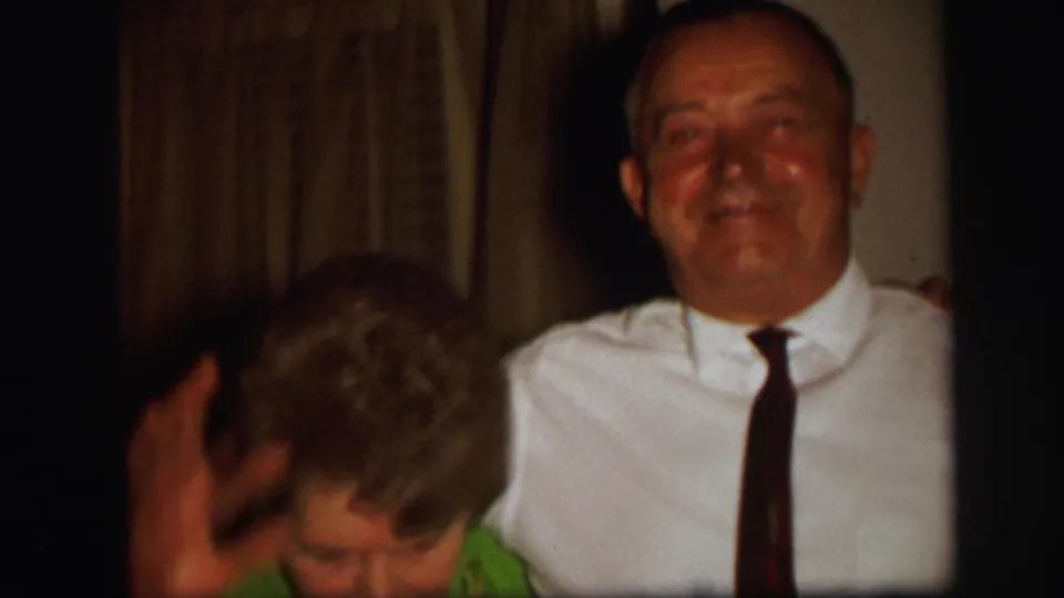 The woman dips her head with a raised hand while the man faces the camera in the 1971 Lynbrook home-movie interior.