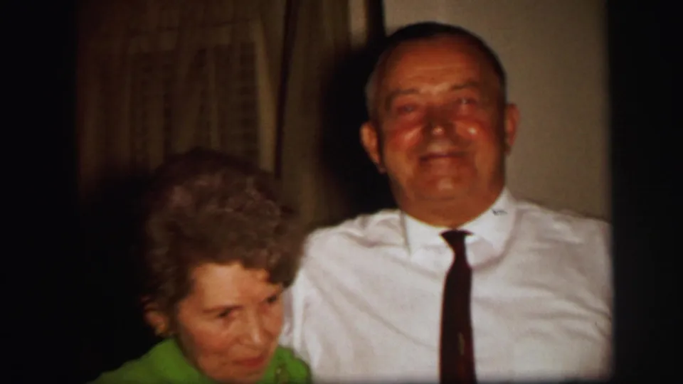 Near the end of the Lynbrook clip, the woman in green leans forward as the man in a white shirt and tie smiles indoors.