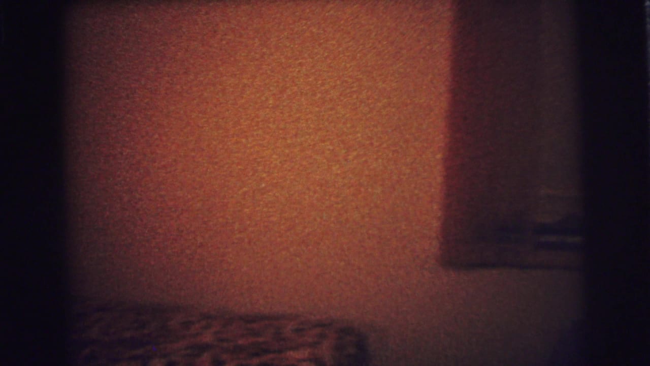 Dim 1973 Lynbrook bedroom frame showing a patterned bed edge, dark wooden headboard, textured wall, and a shadowed wall fixture.