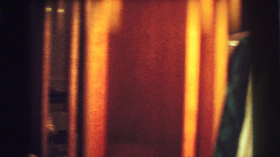 Home-movie frame from the Lynbrook bedroom showing a closet or wardrobe area with hanging clothes, vertical door edges, and warm orange interior light.