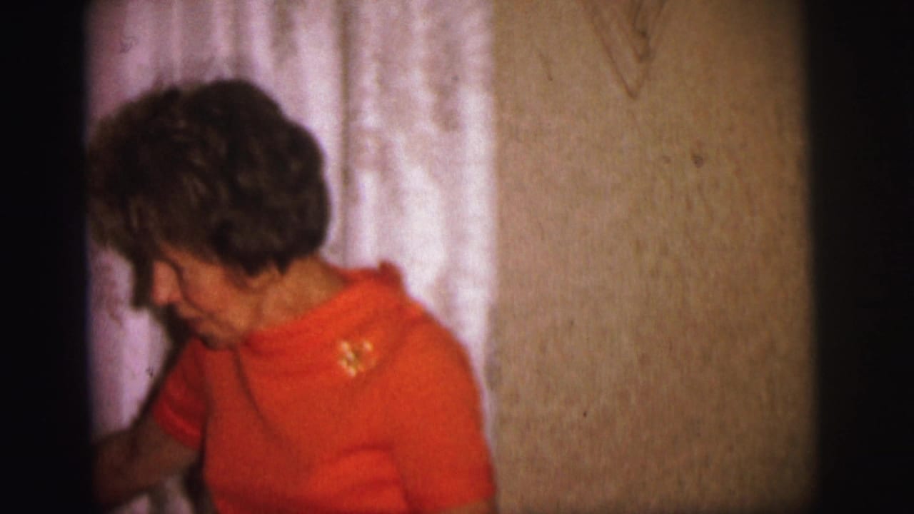 Woman in an orange top stands beside patterned curtains in a dim living room.
