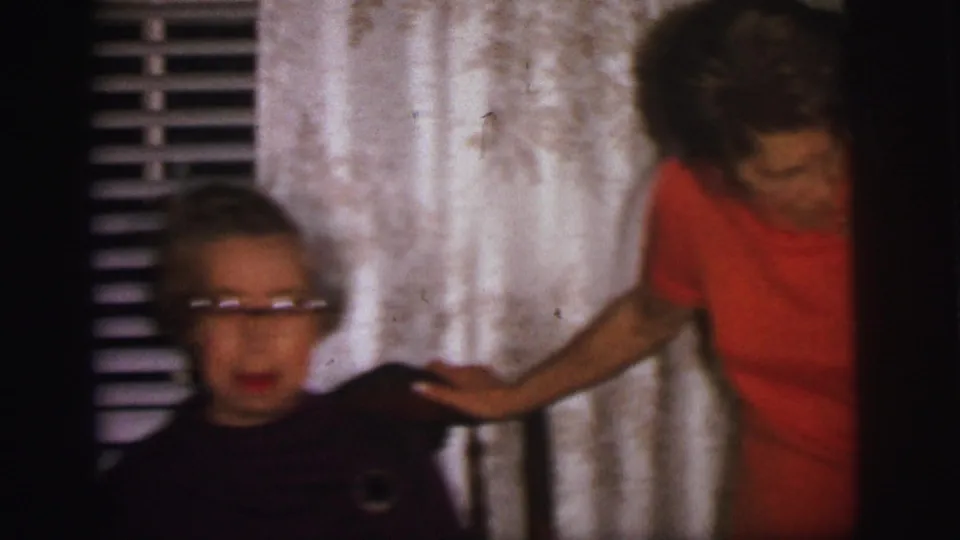 A 1973 Lynbrook home-movie frame shows an older woman with glasses beside curtains as another woman in orange reaches toward her shoulder.