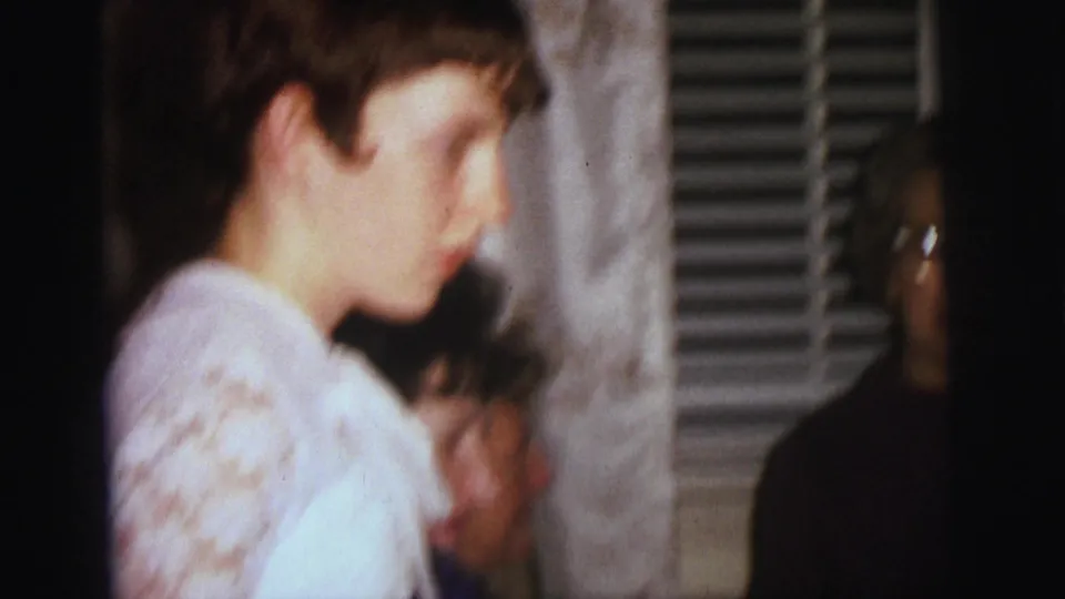 In this Lynbrook 1973 8mm frame, a person in a white lace-trimmed top crosses the foreground with curtains and a partially seen older figure behind.