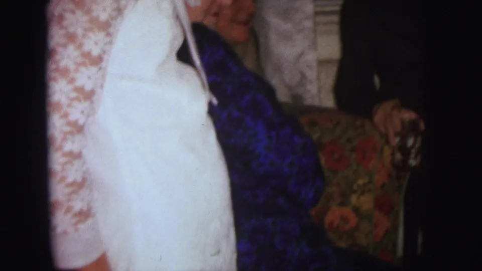 A 1973 Lynbrook home-movie frame shows partially cropped adults near lace curtains, including a bright white jacket and a patterned chair.