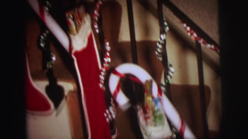 Handheld 1973 home-movie frame of a red Christmas stocking on the stair rail with tinsel, candy cane, and gifts nearby.