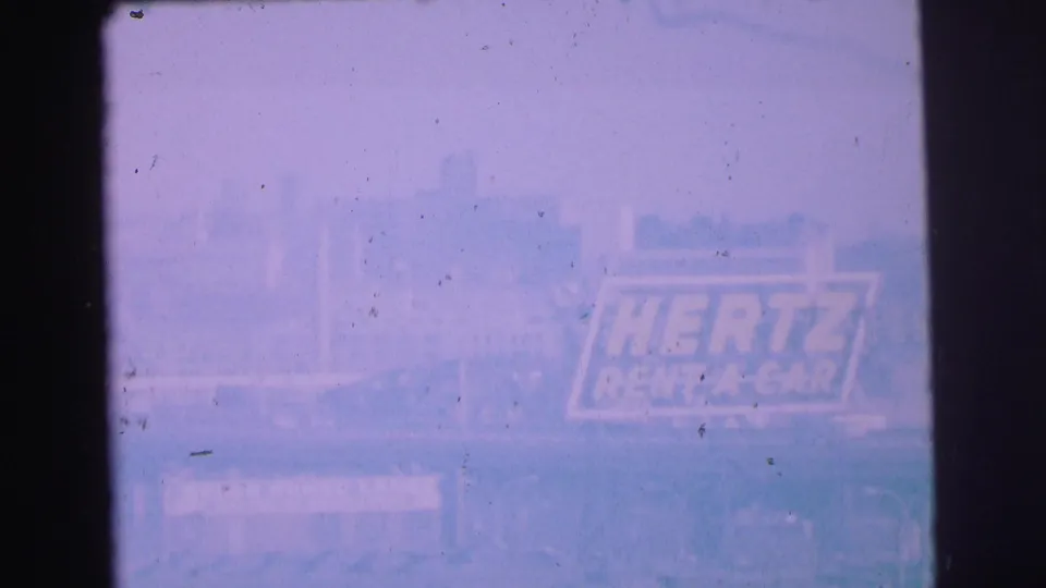 Blurry 1973 Lynbrook frame with washed color, faint roadside structures, and an apparent Hertz rent-a-car sign in the distance.