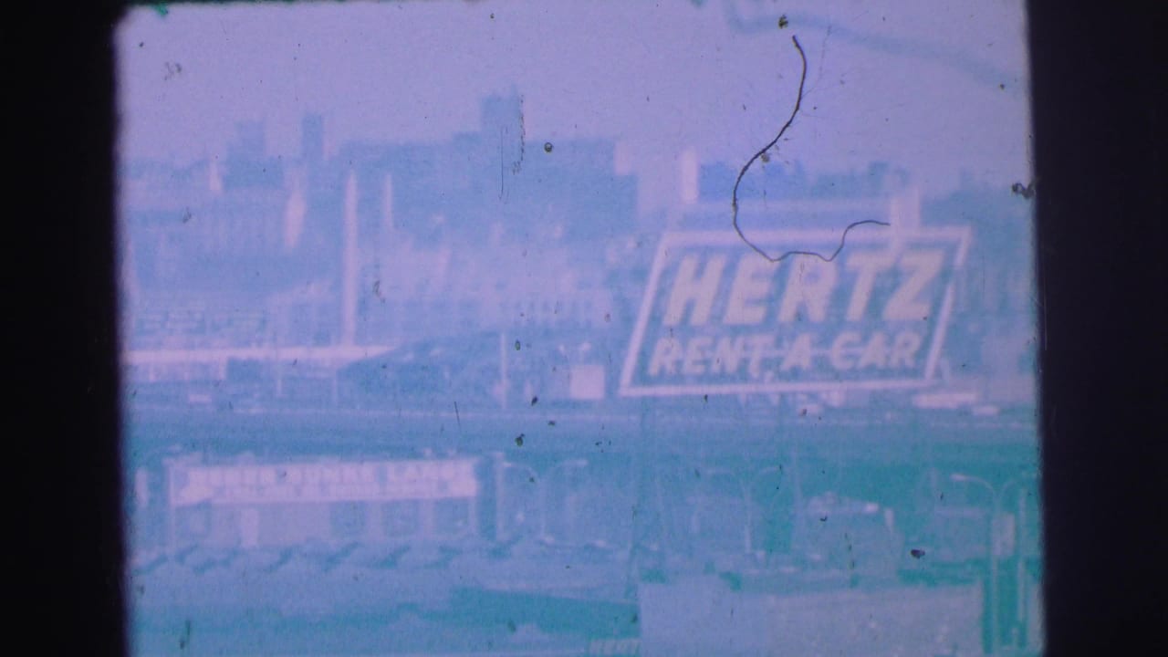 1973 Lynbrook, New York 8mm frame showing a faded Hertz Rent A Car sign through a tinted, weathered moving-vehicle view.
