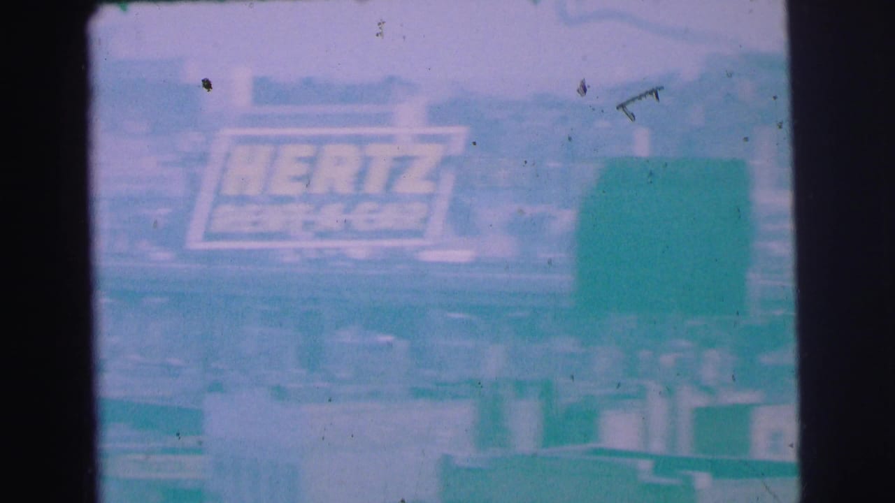 Faded roadside view with the Hertz sign partly visible behind a large greenish shape.