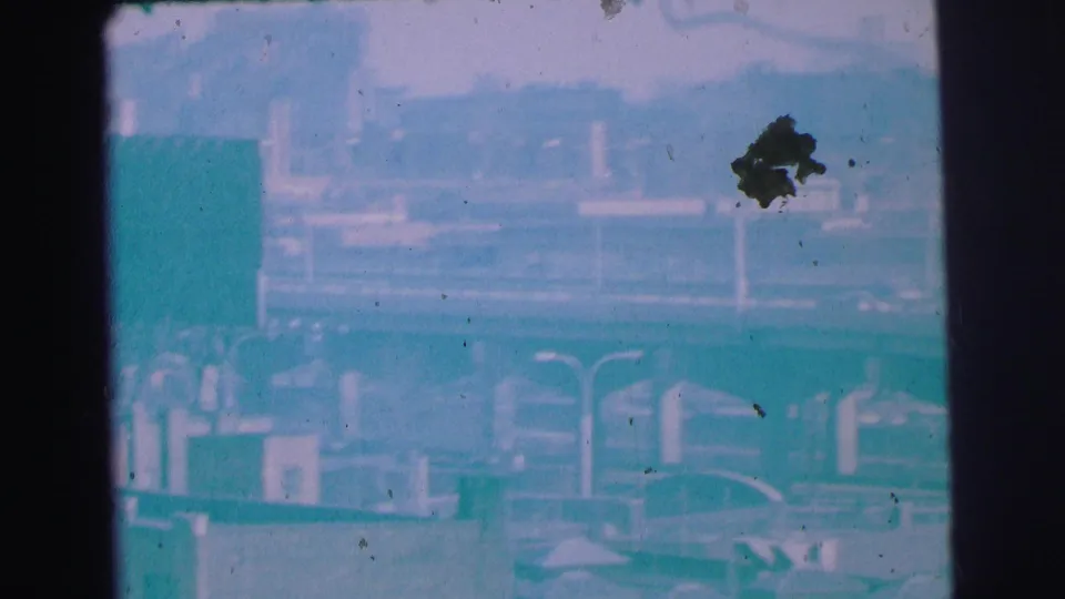 1973 Lynbrook 8mm street frame showing traffic, roadside structures, and a partially obscured commercial area from a moving vehicle.